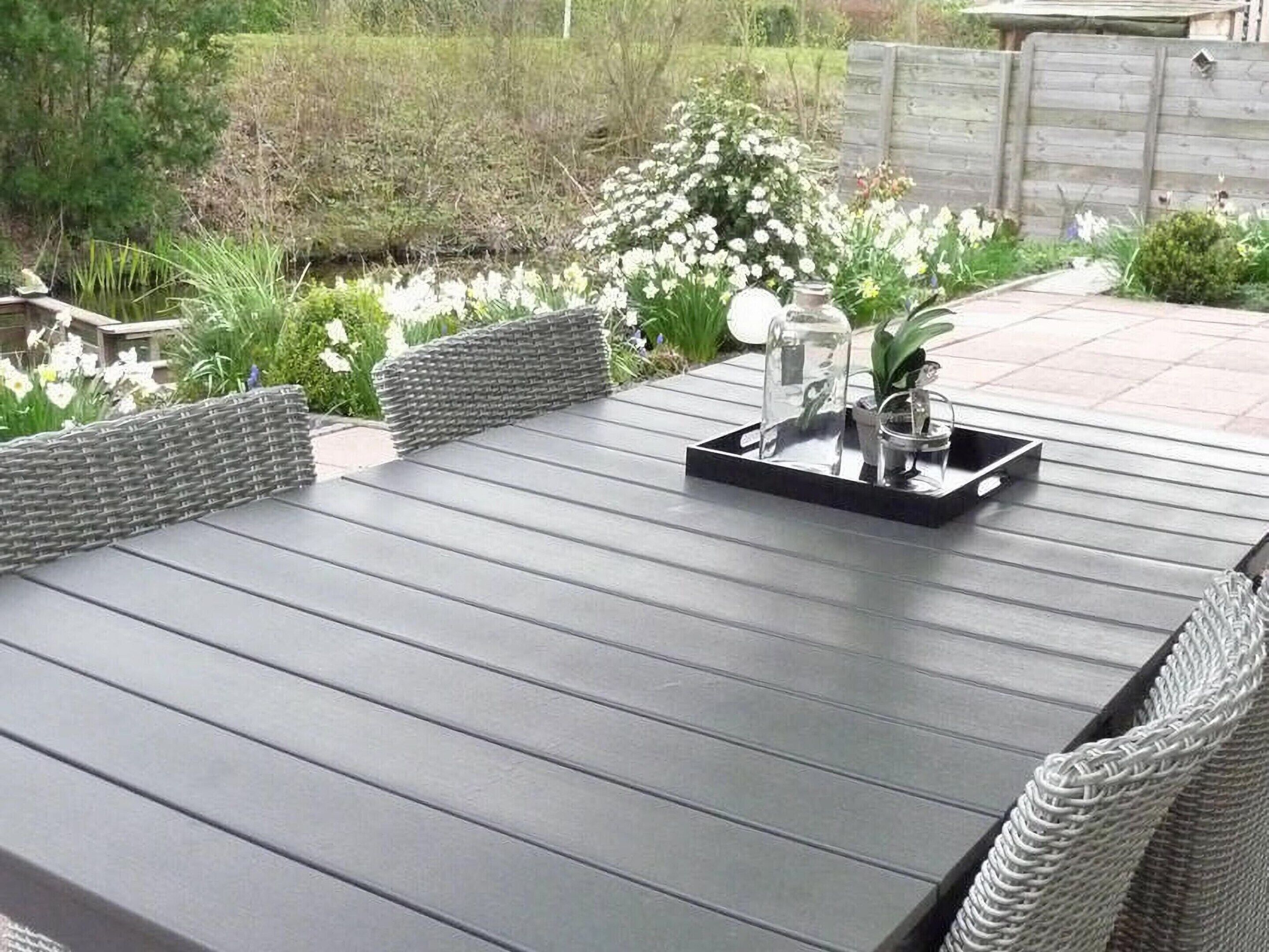 Una mesa de comedor exterior rectangular con acabado oscuro, rodeada de sillas de mimbre. Sobre la mesa, un carrito de vidrio con una botella y un vaso.