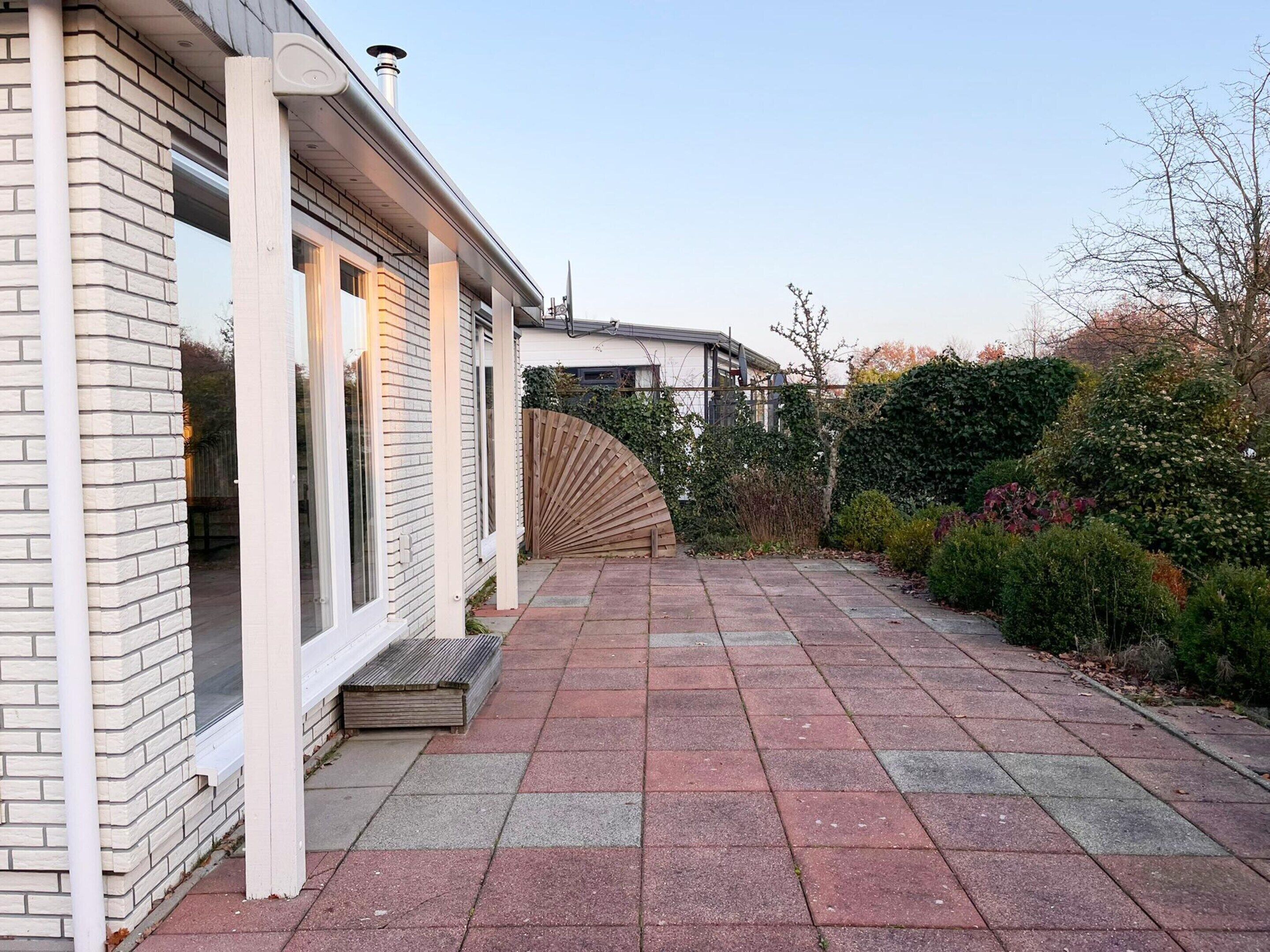 Een bakstenen huis met een overdekte patio, een houten omheining en verschillende struiken en planten.