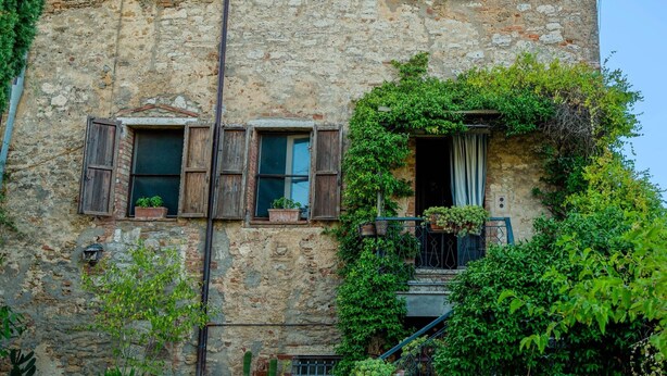Un bâtiment rustique en pierre, doté de volets en bois, d’un balcon orné de plantes en pot et entouré de diverses espèces de verdure.