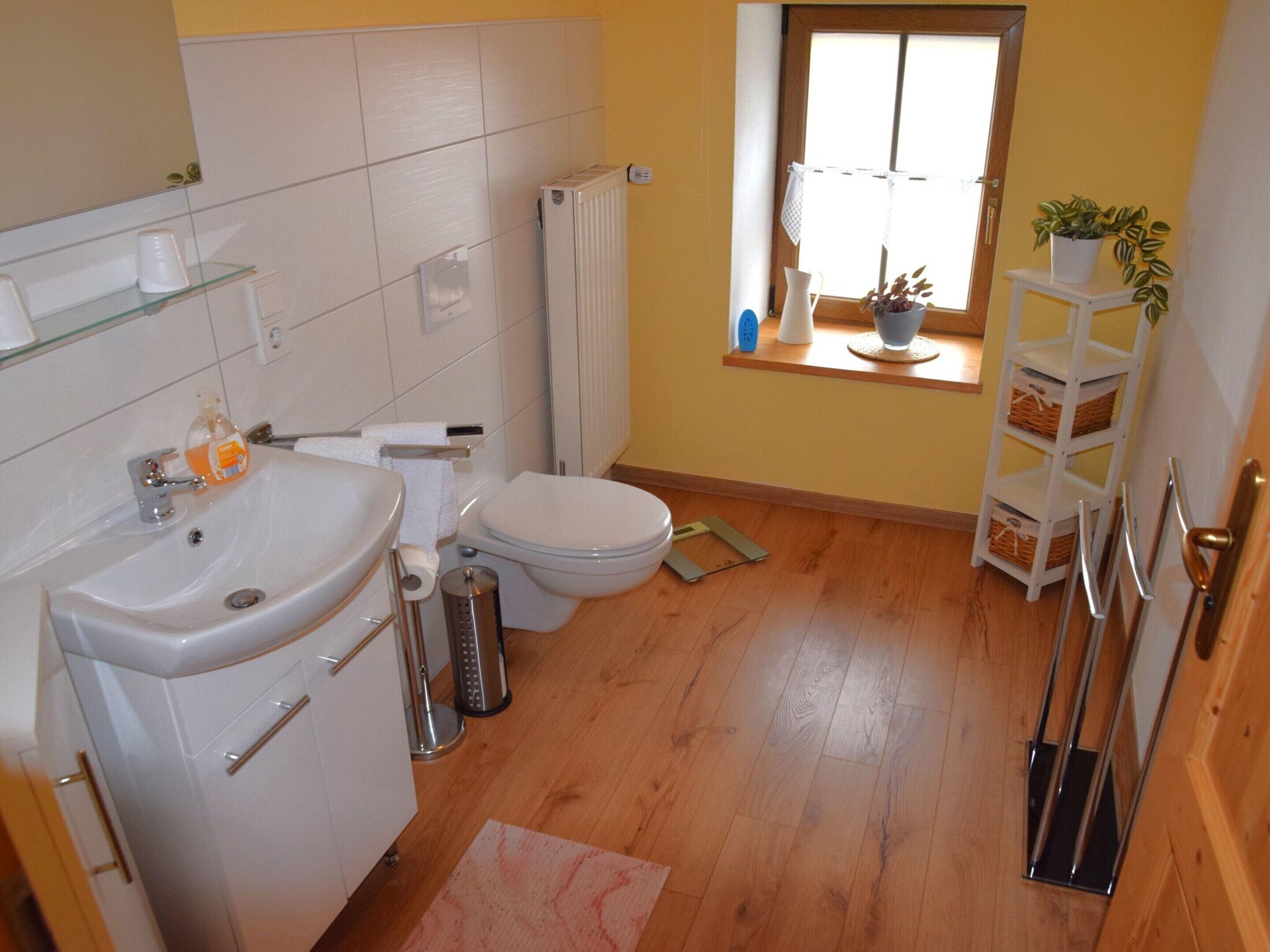A compact bathroom with a white sink, toilet, and radiator. A small window provides natural light, and a shelf holds a plant and a vase.