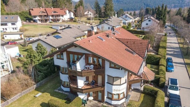 Une maison à deux étages avec un balcon, une cour clôturée et une allée où sont garées des voitures.