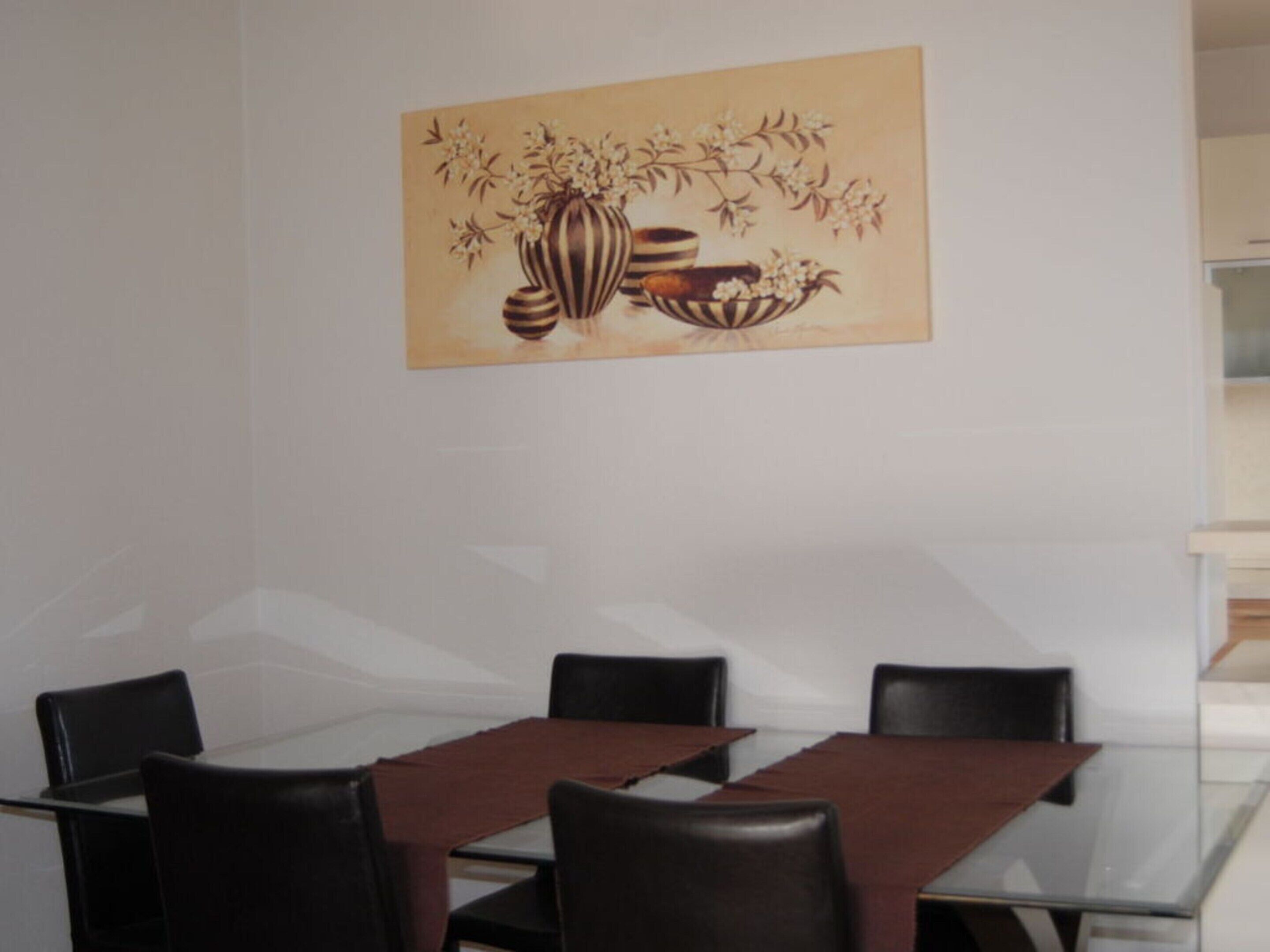 A dining area with a glass table and black chairs, a beige wall with a floral painting, and a white cabinet in the background.