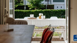 Un área de comedor al aire libre con una mesa preparada para cuatro personas, un florero con flores y vistas a una zona residencial con casas y un árbol.
