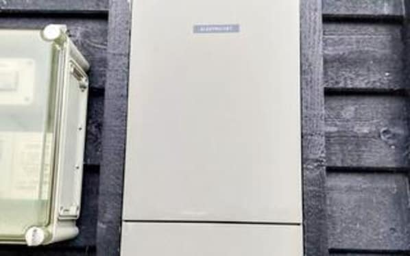 A utility meter with a label, mounted on a wall with a horizontal siding pattern.