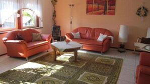 Ein Wohnzimmer mit zwei orangefarbenen Sofas, einem Couchtisch und einem gemusterten Teppich. Es gibt ein Fenster mit Vorhängen, eine Stehlampe und ein Bild an der Wand.