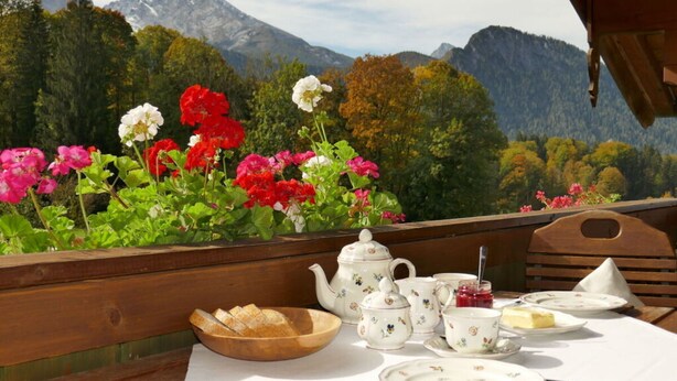 Mesa de madera dispuesta con servicio de té y un cuenco de pan, con vistas a un paisaje montañoso lleno de flores coloridas.