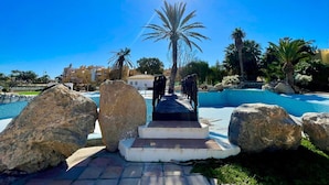 A pool area with a pathway leading to a palm tree, surrounded by large rocks and a clear blue sky. - Apartment in Roquetas Near the Beach (Roquetas de Mar)