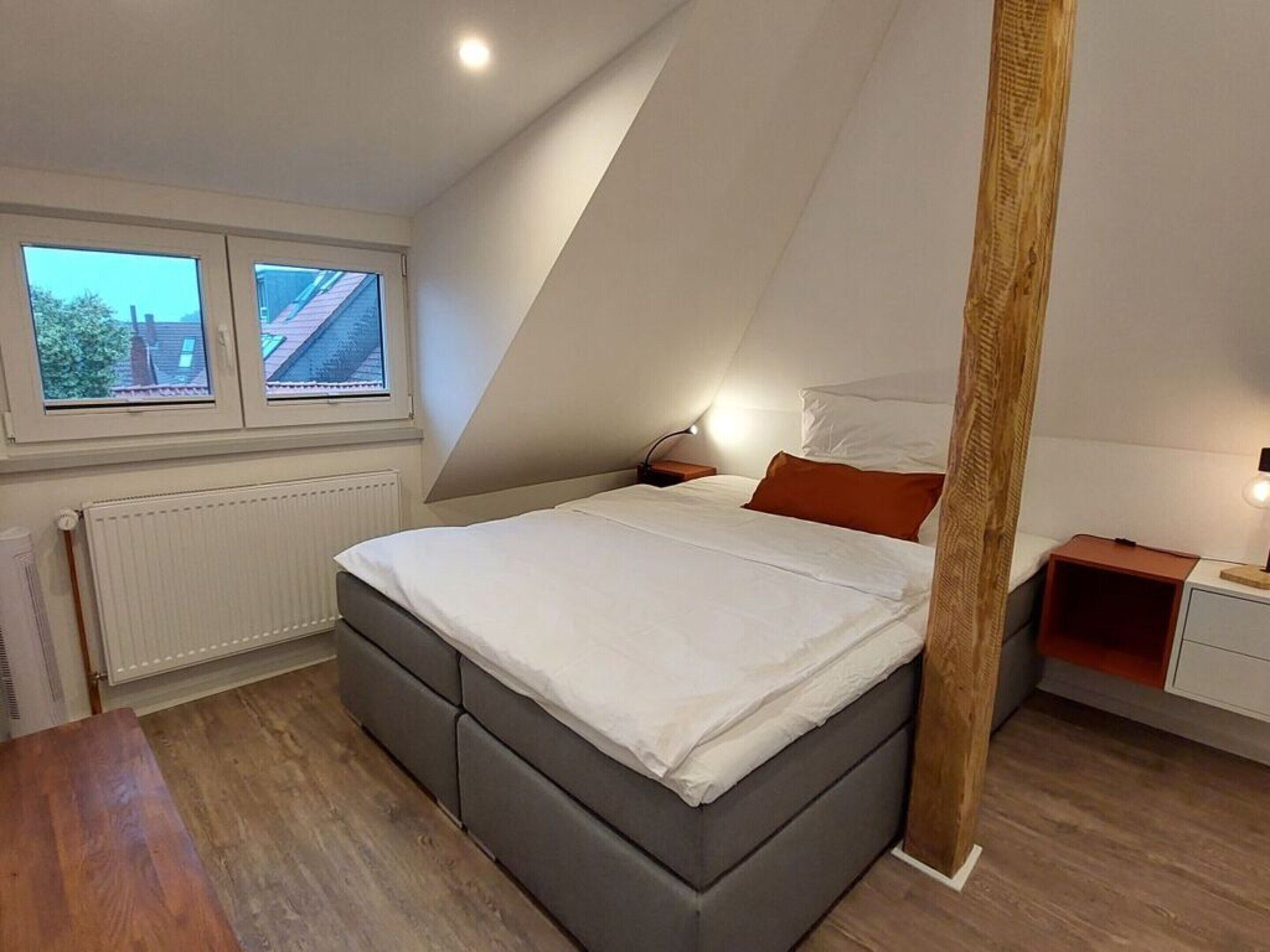 Un dormitorio con techo inclinado, una cama con ropa de cama blanca, una mesita de noche, una silla de madera y una ventana con vistas a una casa.