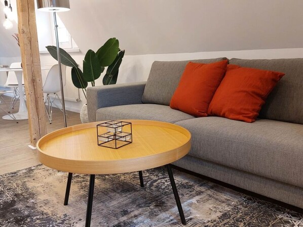 A modern living room with a grey sofa, orange cushions, a wooden coffee table with a geometric glass top, and a large green plant.