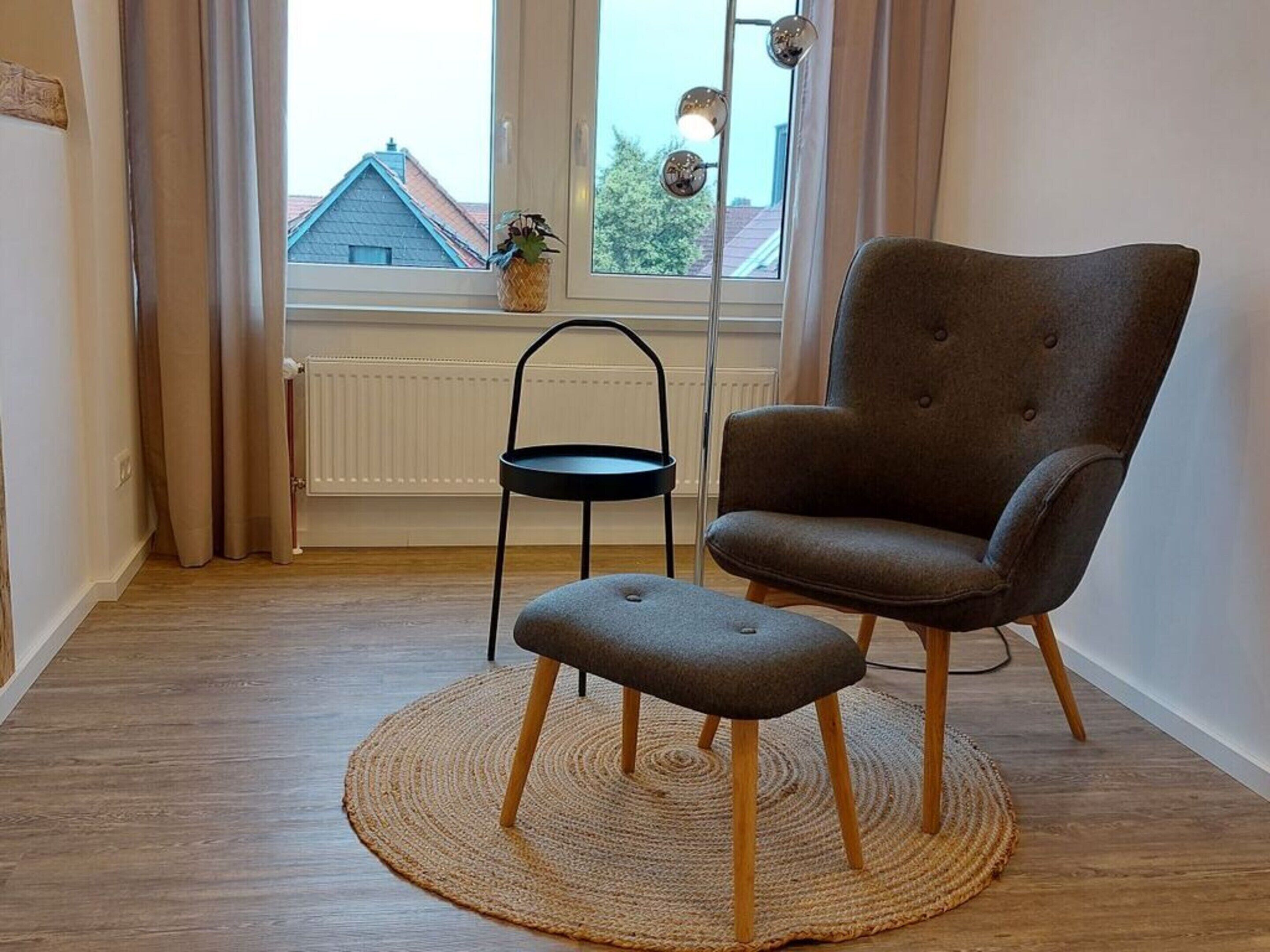 A room with a grey upholstered armchair and a small stool on a woven rug, a black metal chair, a window with curtains, and a radiator.