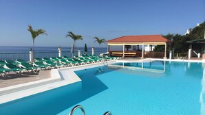 En swimmingpool med liggestole og et overdækket område, omgivet af palmer og fri udsigt til havet.