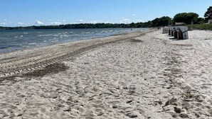 Une plage de sable bordée de cabanes de plage, avec une étendue d’eau calme en arrière-plan.