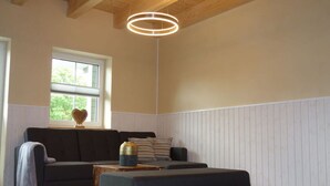 Een moderne woonkamer met een donkere bank, een bijpassend pouf en een houten salontafel. Een bijzondere ronde lamp hangt aan het plafond, en een raam zorgt voor natuurlijk licht.