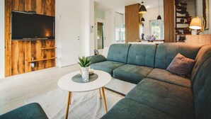 A modern living room with a sectional sofa, a flat-screen TV mounted on a wooden wall, a round coffee table with a plant, and a dining area with a lamp. - La Mia Storia With Hot tub (Beretinec)