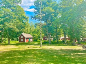 En röd trästuga med en grön dörr, omgiven av frodiga, gröna träd och gräs.