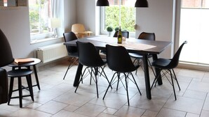 Un coin repas avec une table rectangulaire, six chaises noires, deux lampes suspendues, une horloge au mur et une fenêtre avec des rideaux.
