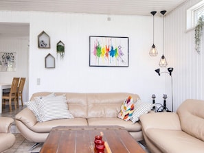 A living room with a beige sofa, a wooden coffee table, and a colorful abstract painting. There are two hanging pendant lights and a small plant on the wall.