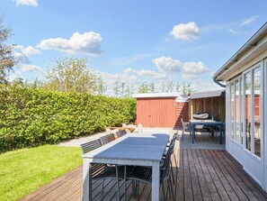 Une terrasse extérieure avec une table et des chaises, une terrasse en bois, un abri de jardin rouge et une haie.