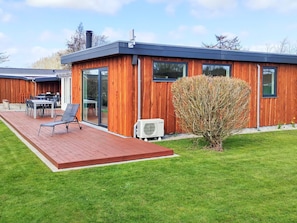 A modern wooden house with a deck, outdoor furniture, and a small tree.