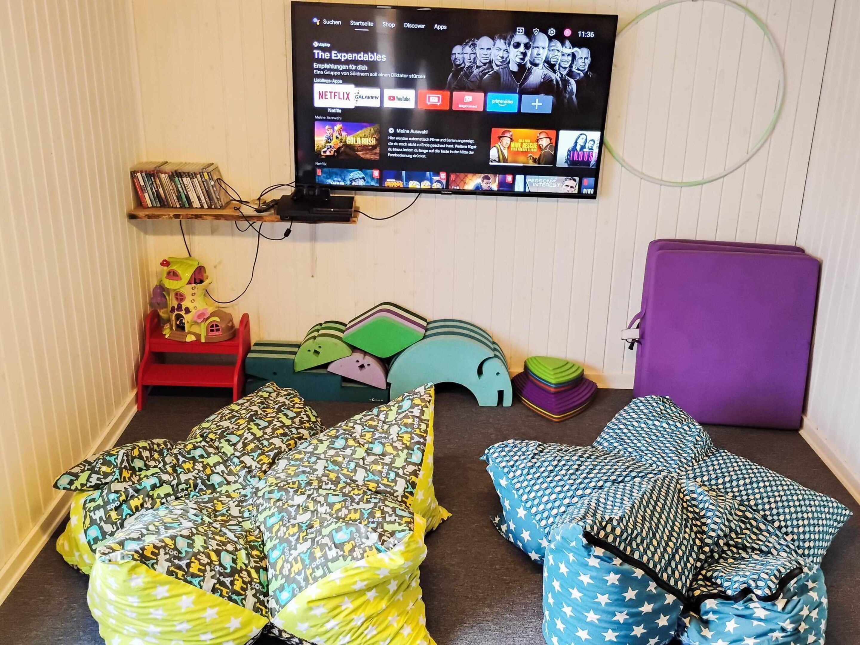 A room with a TV displaying a streaming service interface, a small bookshelf, and two beds with patterned bedding.