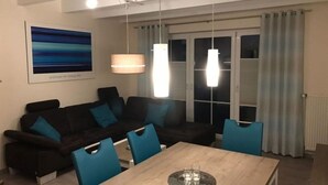 A modern living room with a dark sofa, blue cushions, a wooden dining table, and a large window with curtains. - Seepark Burhave Haus 8 (Butjadingen)