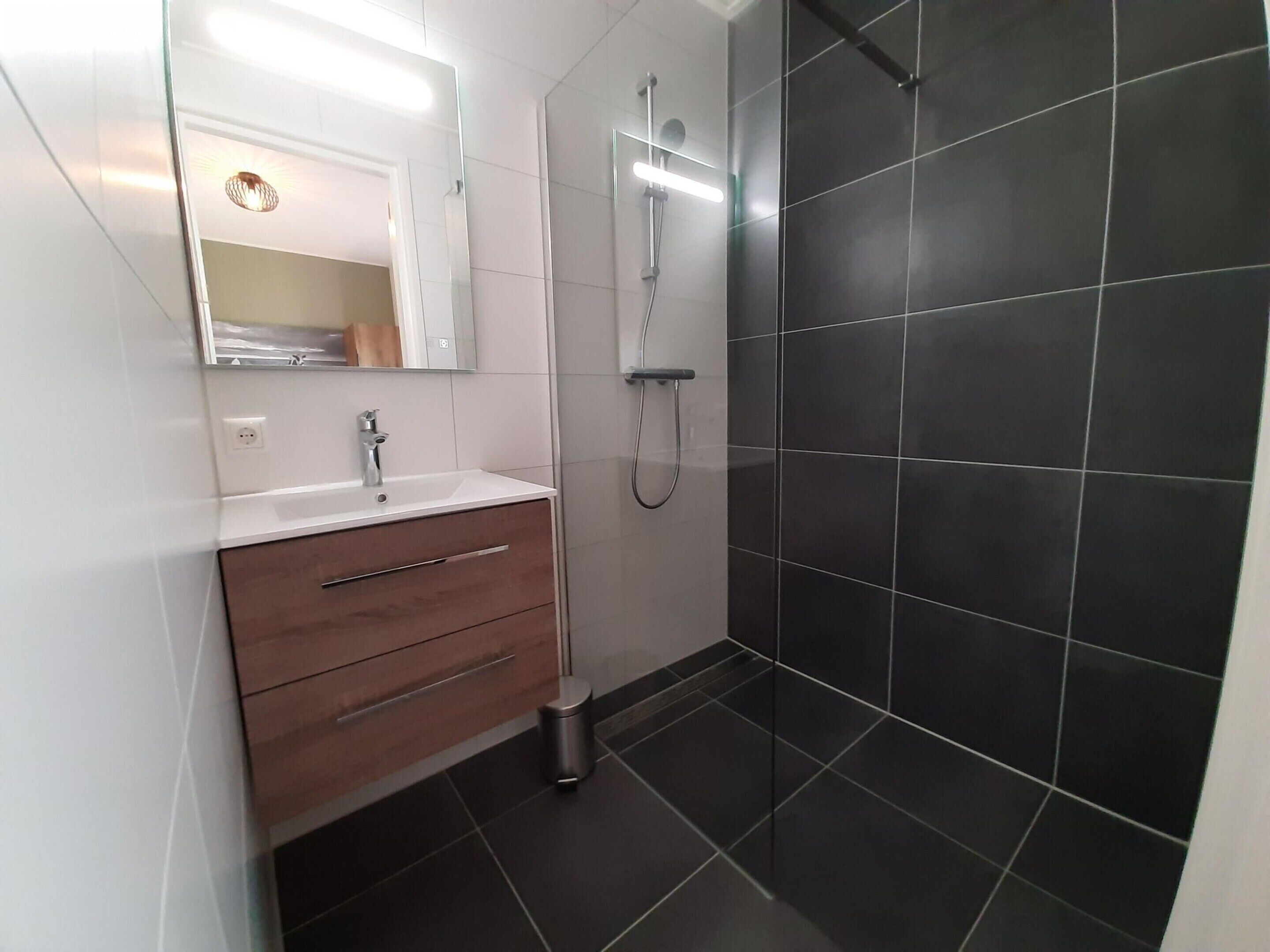 Ein modernes Badezimmer mit einem weißen Waschtisch, einem Spiegel, einer Dusche inklusive Handbrause und dunkel gefliesten Wänden.