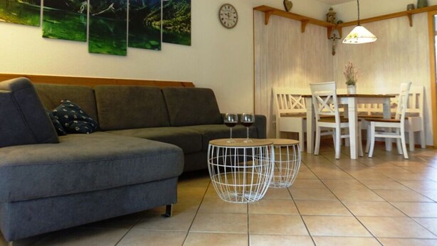 A living room with a grey sofa, a dining table with white chairs, and two round wire tables with wine glasses. - Sandra Comfortable Holiday Residence (Lechbruck)