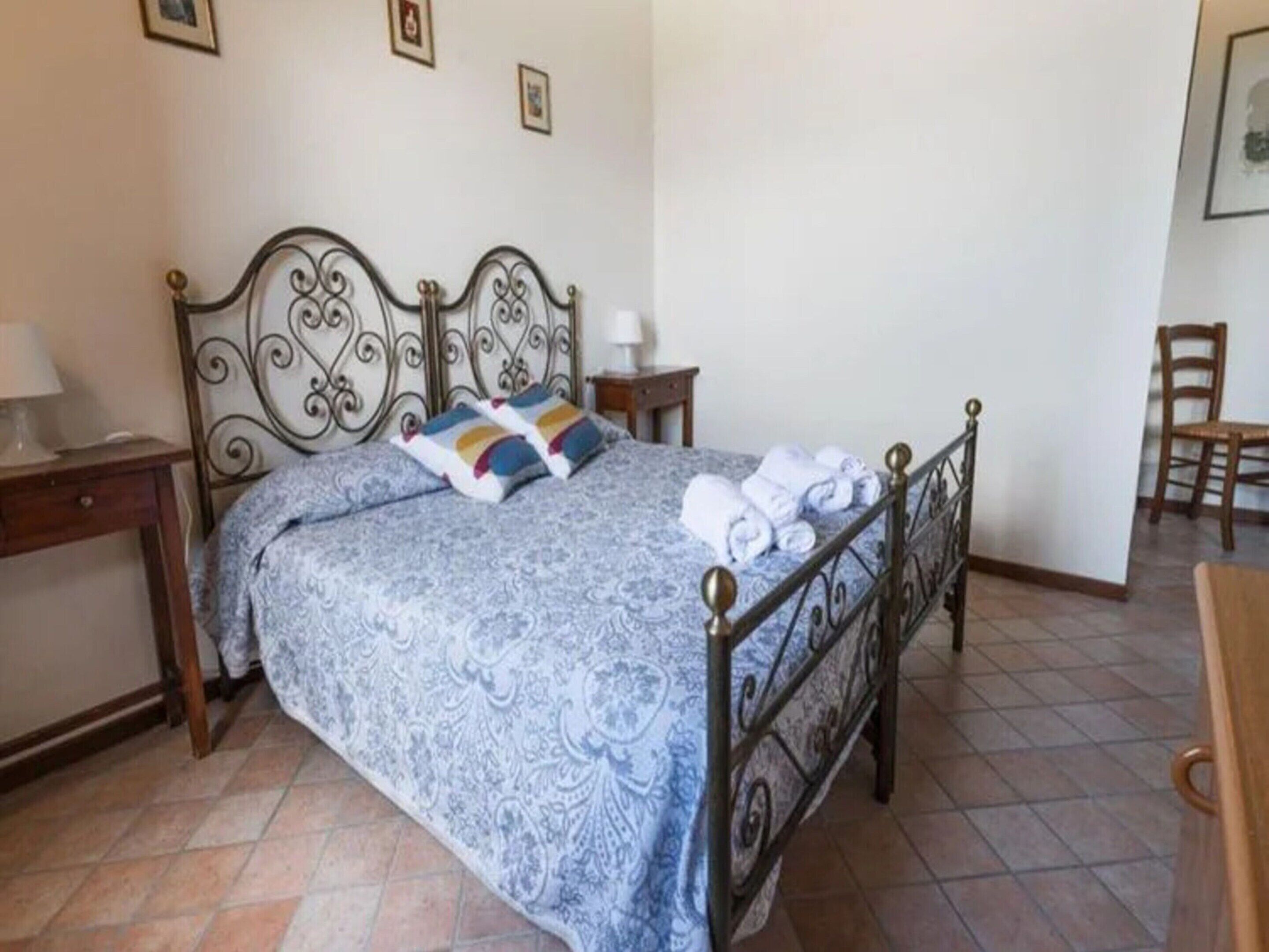 Un lit bien fait, avec une couvre-lit à motifs et des oreillers, une table de chevet avec une lampe, et une chaise dans un coin.