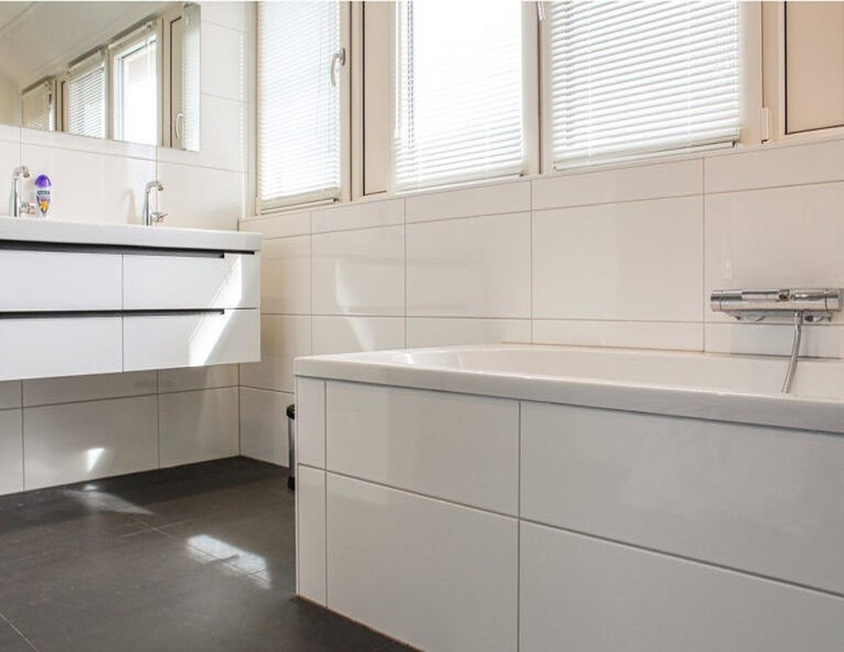 Een moderne badkamer met een wit wastafelmeubel, een bad en een raam met luxaflex.