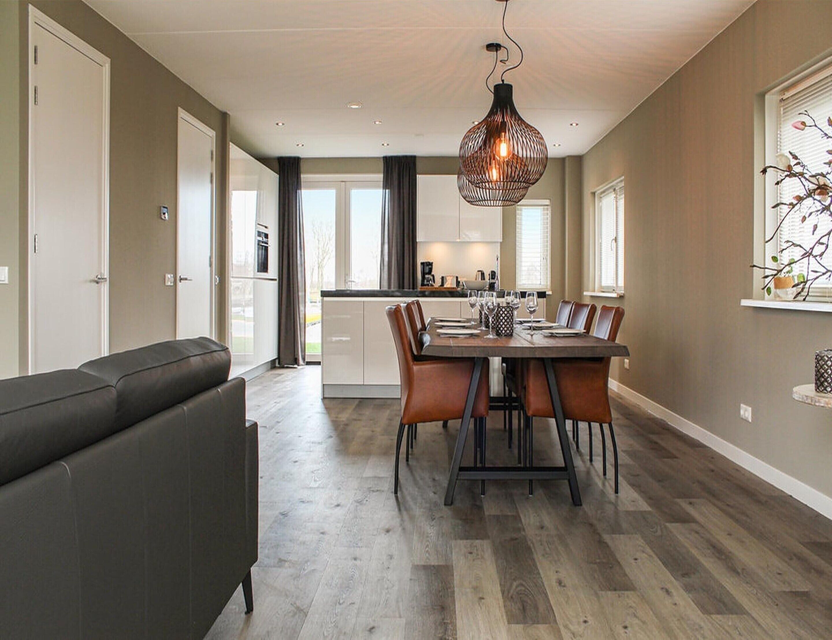 A modern kitchen with a dining area, featuring a wooden table and chairs, a hanging light fixture, and a view of the outdoors.