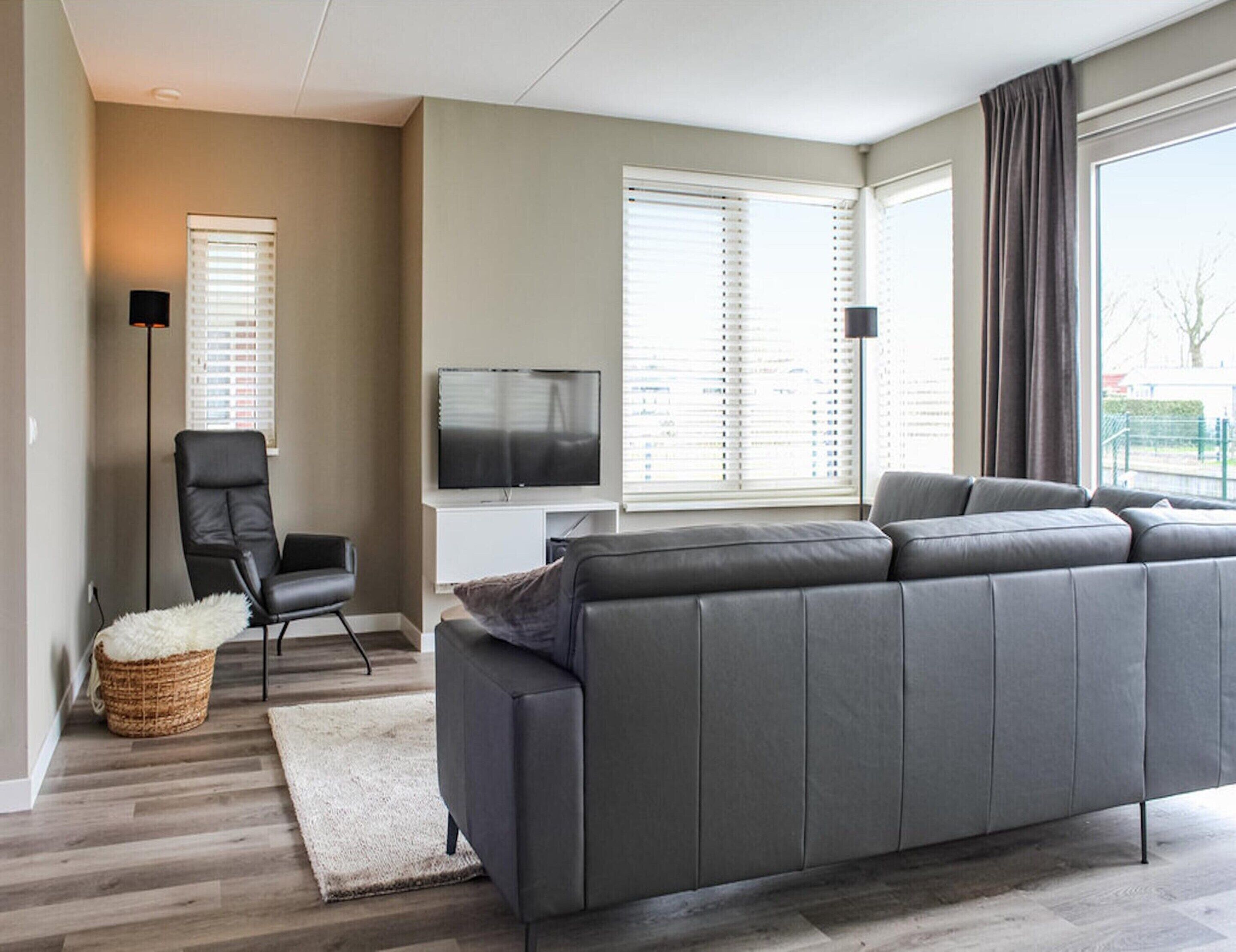 A modern living room with a grey sofa, a black chair, a TV, and a large window with blinds.