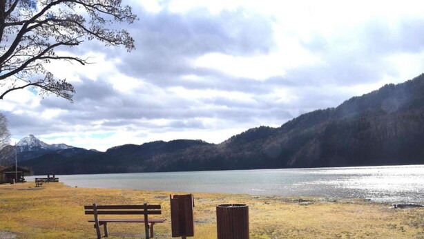 A lakeside park with a bench and trash can, surrounded by mountains and a cloudy sky. - Family Escape With Pool (Nesselwang)
