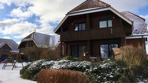 A two-story wooden house with a balcony, surrounded by snow-covered bushes and a clear sky. - Small Sundblick by the sea in Lemkenhafen (Fehmarn)