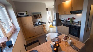 A modern kitchen with wooden cabinets, a microwave, a stove, and a dining area with a table set for two. - Small Sundblick by the sea in Lemkenhafen (Fehmarn)