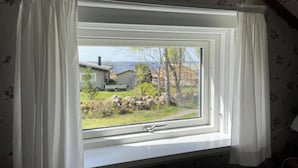 Una habitación con ventana que da a un paisaje rural, con cortina blanca, cama y papel tapiz floral.