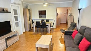 Ett modernt vardagsrum med en platt-TV, en soffa med röda kuddar, en matplats med bord och stolar samt ett kök i bakgrunden.