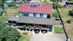 A two-story building with a red roof, a covered porch with potted plants, and a paved driveway. - Charming Apartment by Worthersee (Techelsberg am Worther See)