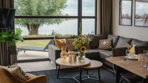 Uma sala de estar moderna com uma janela ampla com vista para um lago, um sofá, uma mesa de centro e uma área de jantar.