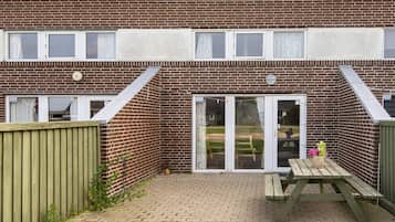 Edificio de ladrillo con un patio que incluye una mesa y sillas de madera, un jardín cercado y una puerta corrediza de vidrio que da a un espacio exterior cubierto.