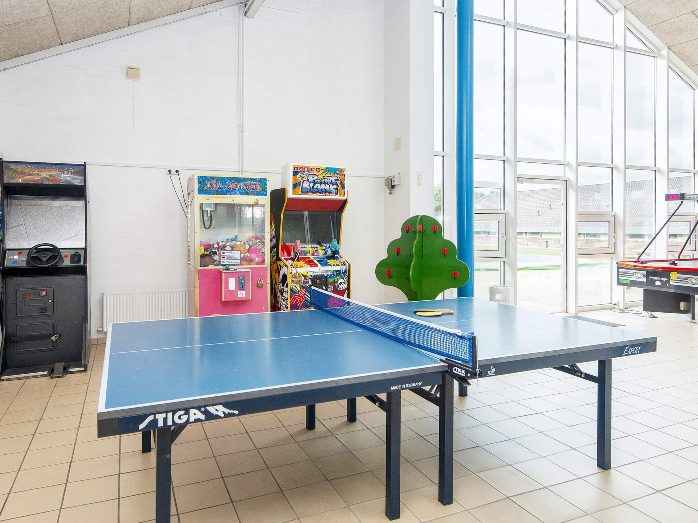 A ping pong table with a blue net, surrounded by arcade games and a large window.