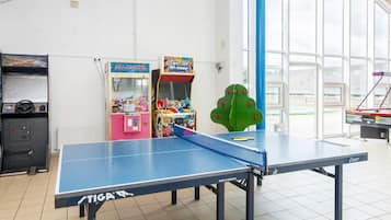 A ping pong table with a blue net, surrounded by arcade games and a large window.