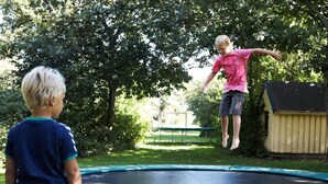 Ein Trampolin in einem Garten mit einer Holzhütte und Bäumen im Hintergrund.