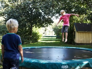 Ein Trampolin in einem Garten mit einer Holzhütte und Bäumen im Hintergrund.