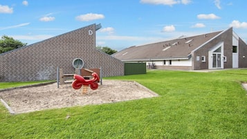A playground with a red scooter and a sandpit, a brick wall with a 'D' sign, and a building with a sloped roof.