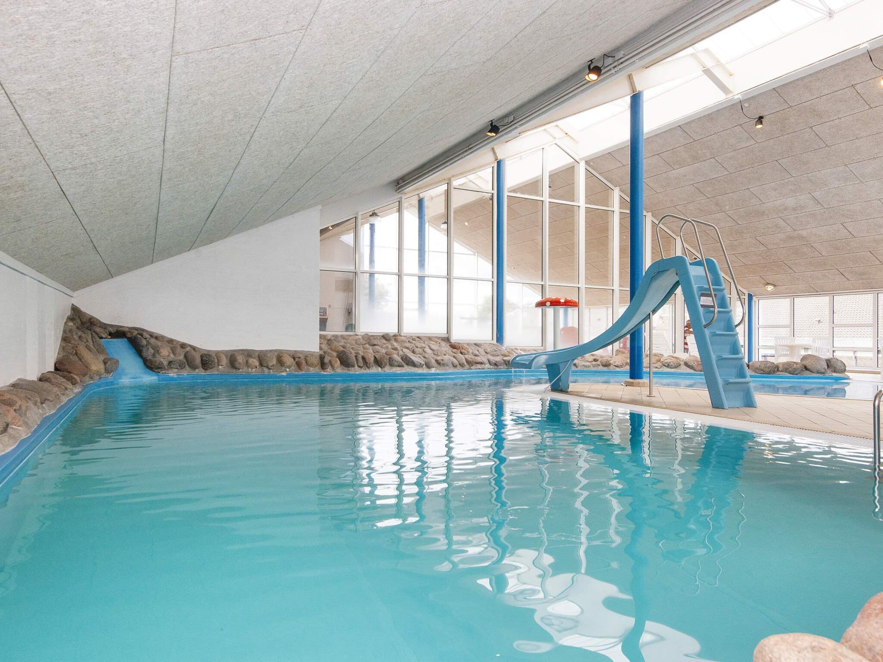 An indoor swimming pool with a blue slide, stone walls, and large windows.