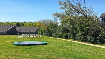 A grassy outdoor area with a trampoline, a building with a sloped roof, and a pathway leading to it.