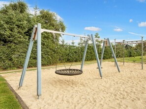 Une aire de jeux avec des balançoires classiques et une balançoire en forme de pneu, entourée de sable et de verdure.