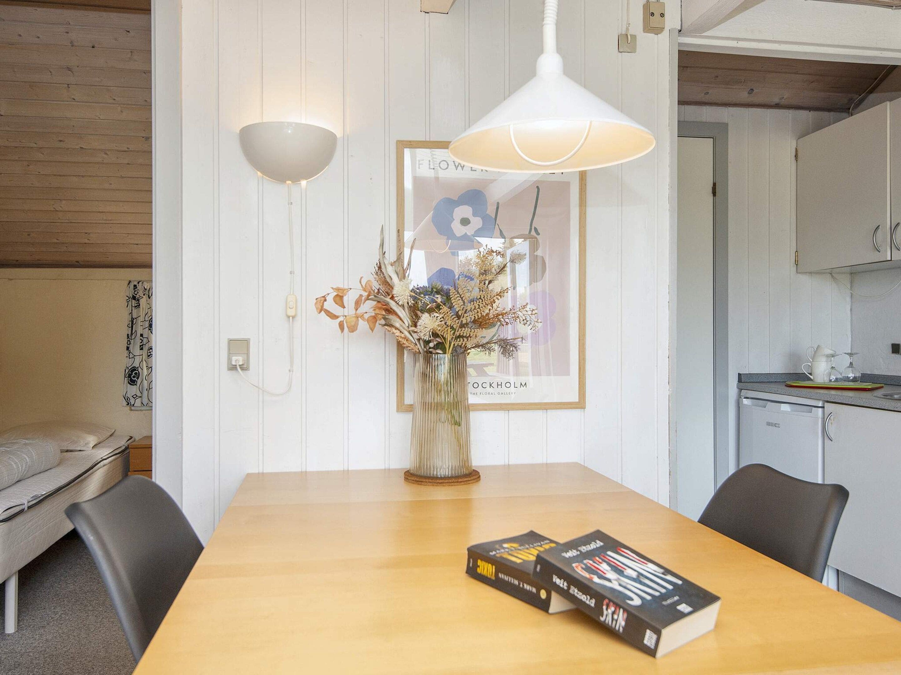 A dining area with a wooden table, two chairs, a vase with dried flowers, and two books.