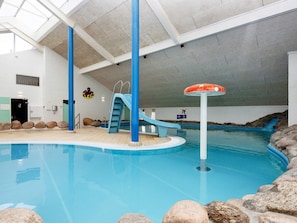 Une piscine intérieure avec un toboggan bleu, un poste de surveillance pour les sauveteurs et des bordures de piscine recouvertes de pierres.