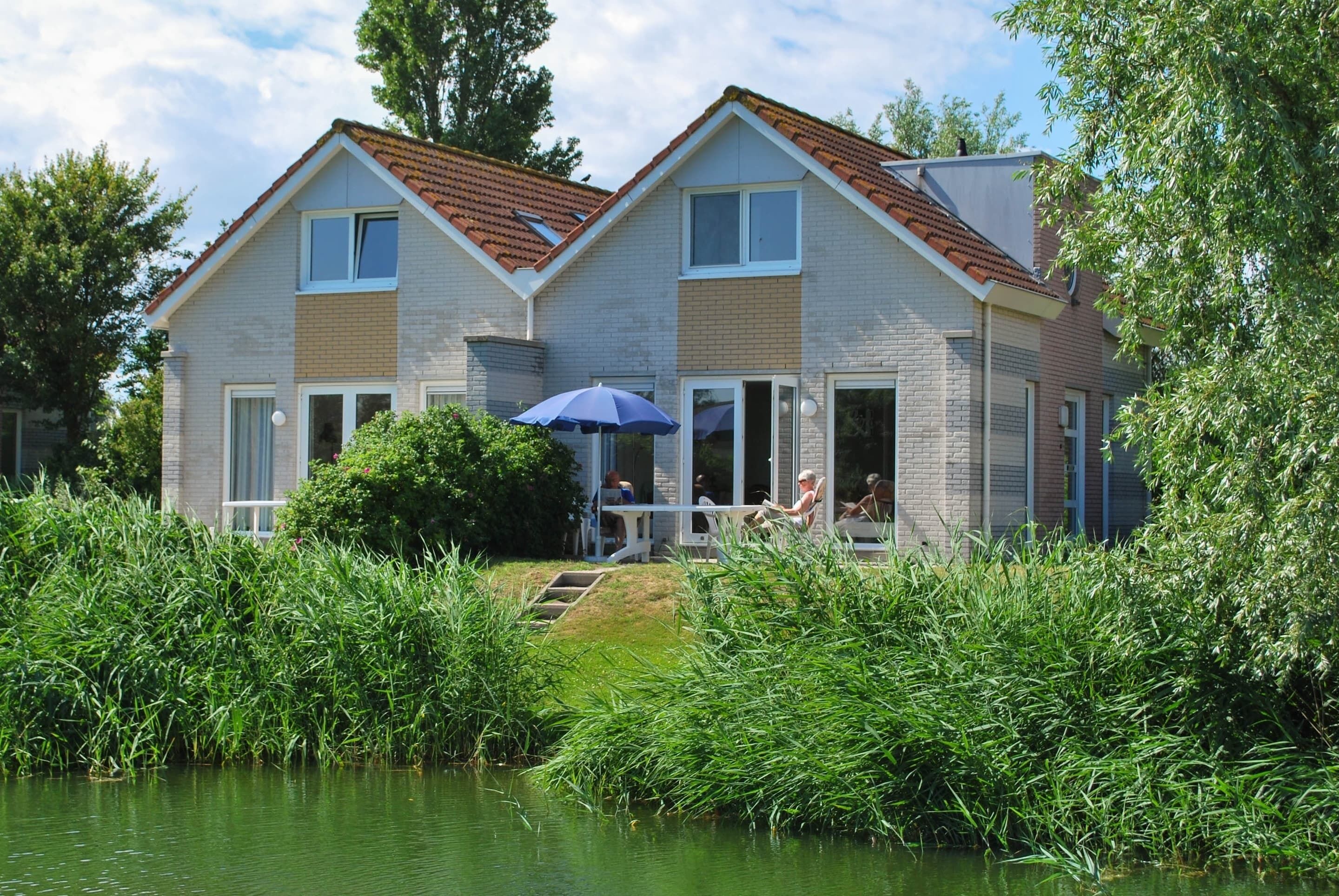 Een huis met twee verdiepingen, voorzien van een dak met rode tegels, een overdekte veranda en een vijver op de voorgrond.